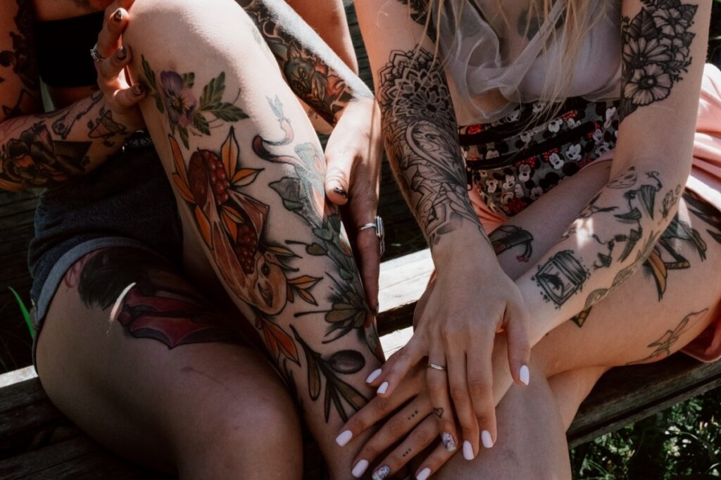 Two female sit on a bench, showing their tattoo art