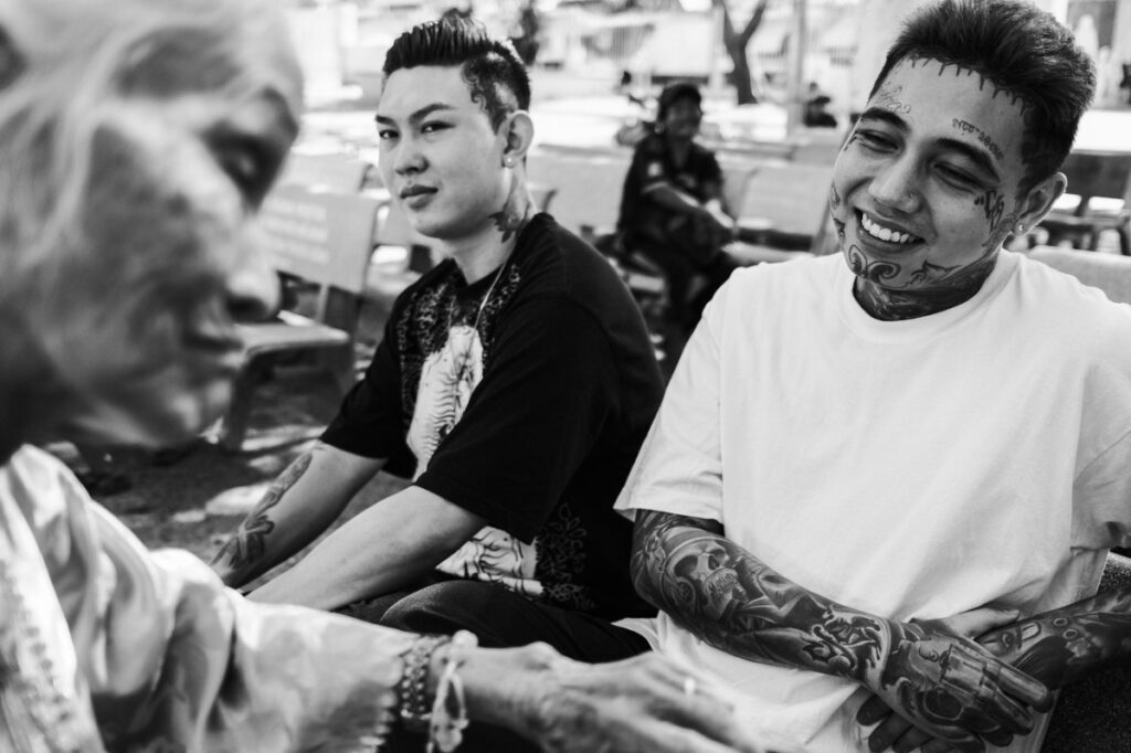 A man sits in a tatoo shop, ink of his arms, smiling at an older lady