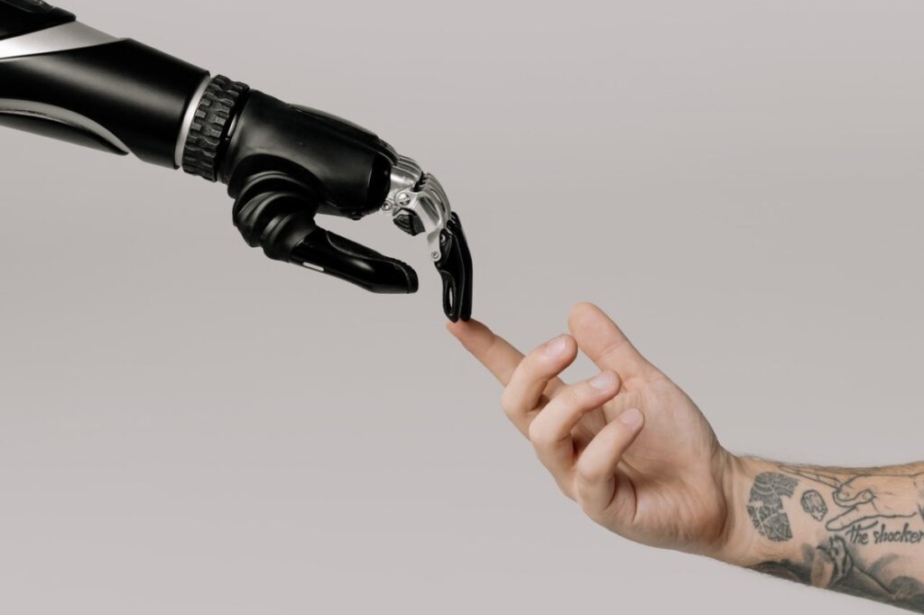 A robotic hand is outstretched, touching the finger tips of a real tattooed hand and arm